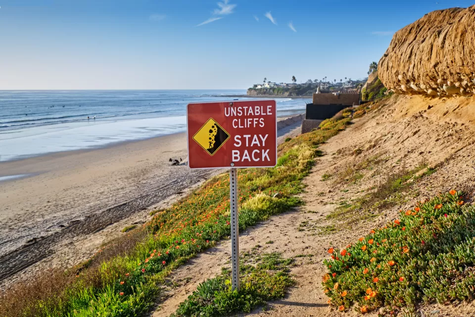 Red "Unstable Cliffs - Stay Back" sign on tall Pacific coast cliff